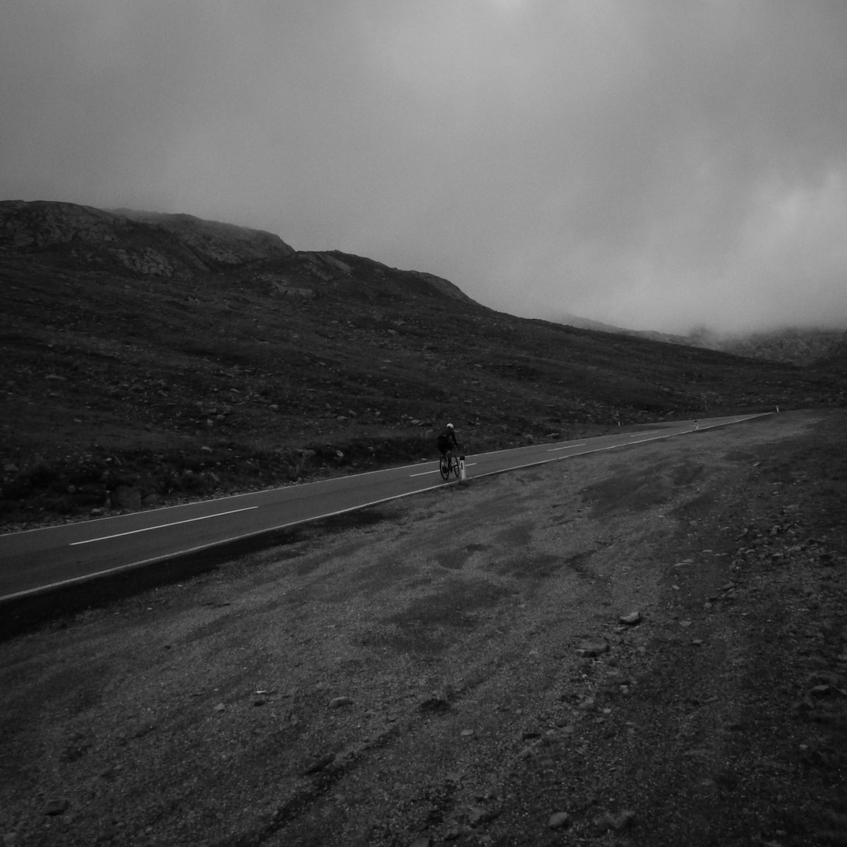 A person cycling on a road through a misty mountain landscape in black and white