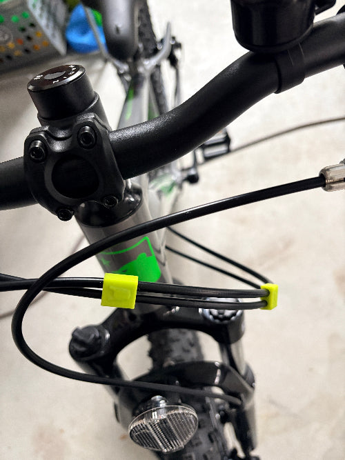 Close-up of bicycle handlebars and front frame with attached cables secured by two Brake and Shift Cable Clips (pack of 5). A headlight is mounted on the front fork, with a blurred indoor background.