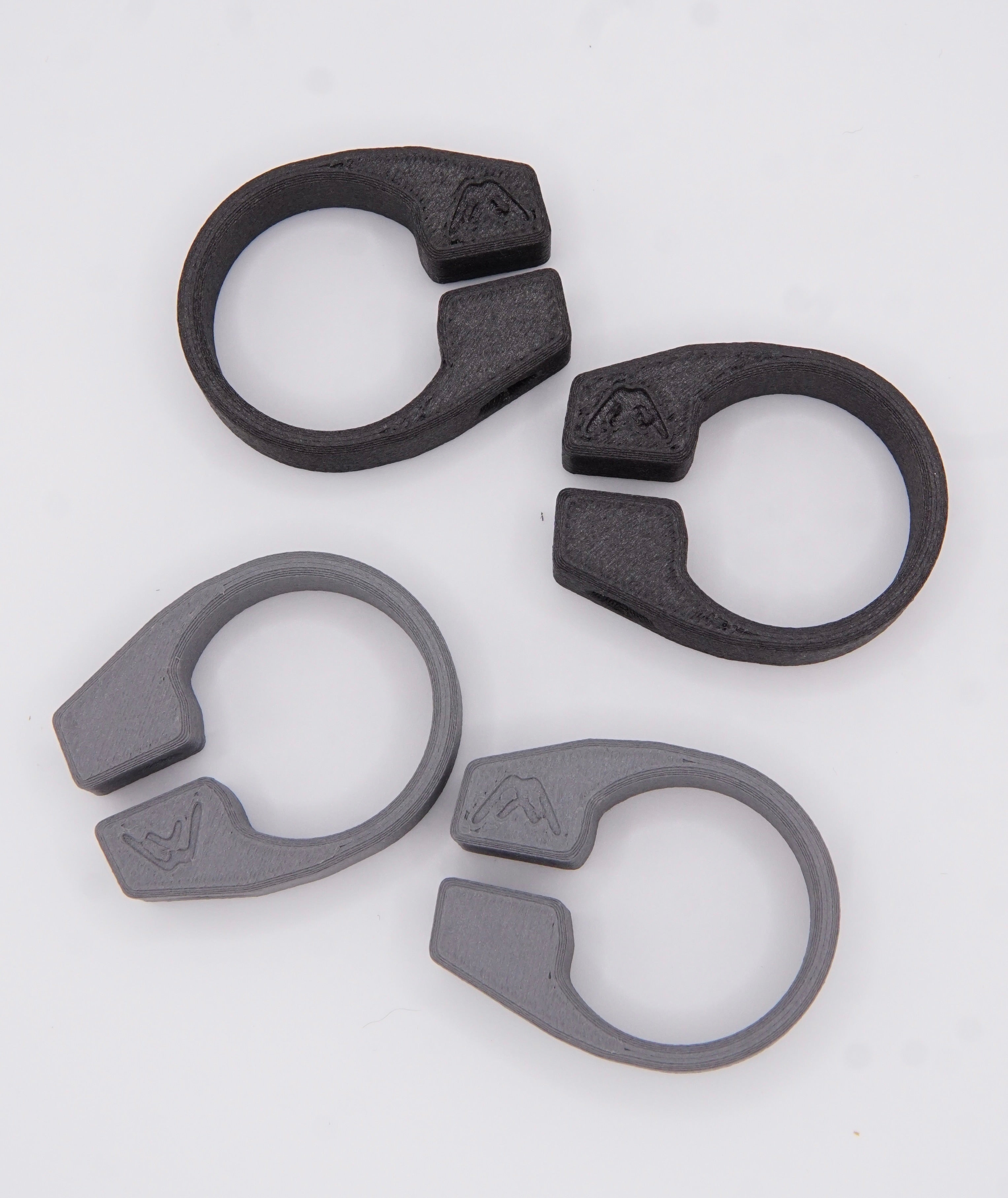 Four MTB AirGrips Spare Clamps—two black and two gray ring-shaped plastic clips—are arranged on a white background. Each interchangeable clamp, featuring a small mountain symbol, fits as replacement hardware for AirGrip bar clamps.