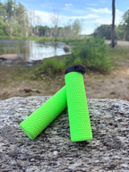 Two MTB Air Grips - Green Monstah! with a textured, bright green design rest on a large rock, overlooking a calm lake, trees, and a partly cloudy sky.
