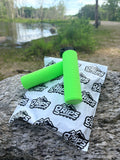 Two bright green MTB Air Grips - Green Monstah! rest on a large rock beside a white TAN Grips package, with a river and trees visible in the background.