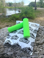 Two bright green MTB Air Grips - Green Monstah! rest on a large rock beside a white TAN Grips package, with a river and trees visible in the background.