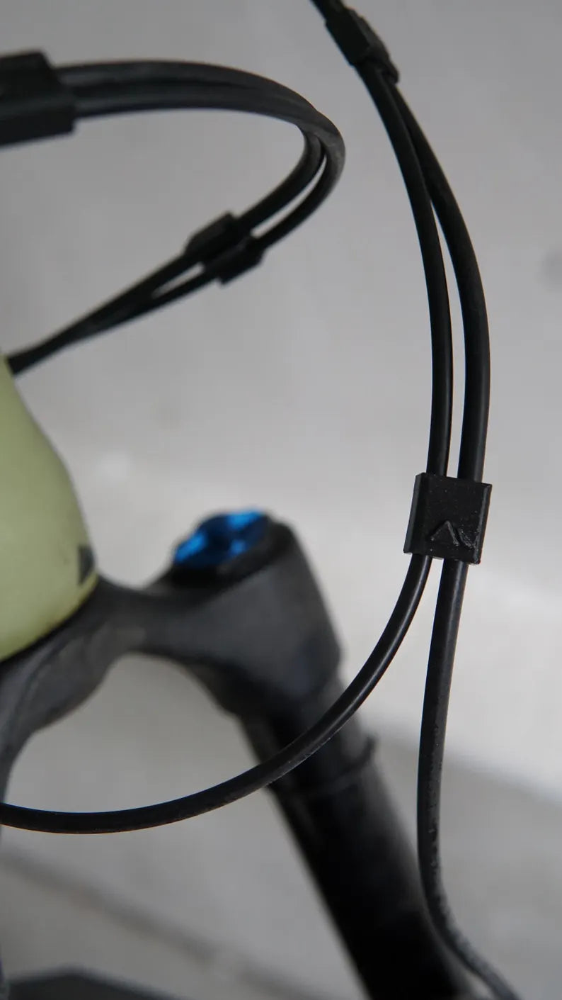 Close-up of black bicycle cables secured with a Brake and Shift Cable Clip (pack of 5), with part of the bike frame and a blue bolt visible in the background.