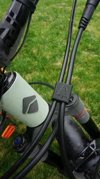 Close-up of a light green mountain bike’s front cables and handlebars with one of our Brake and Shift Cable Clips (pack of 5), featuring a small rubber piece with a pine tree symbol. Green grass is visible in the background.